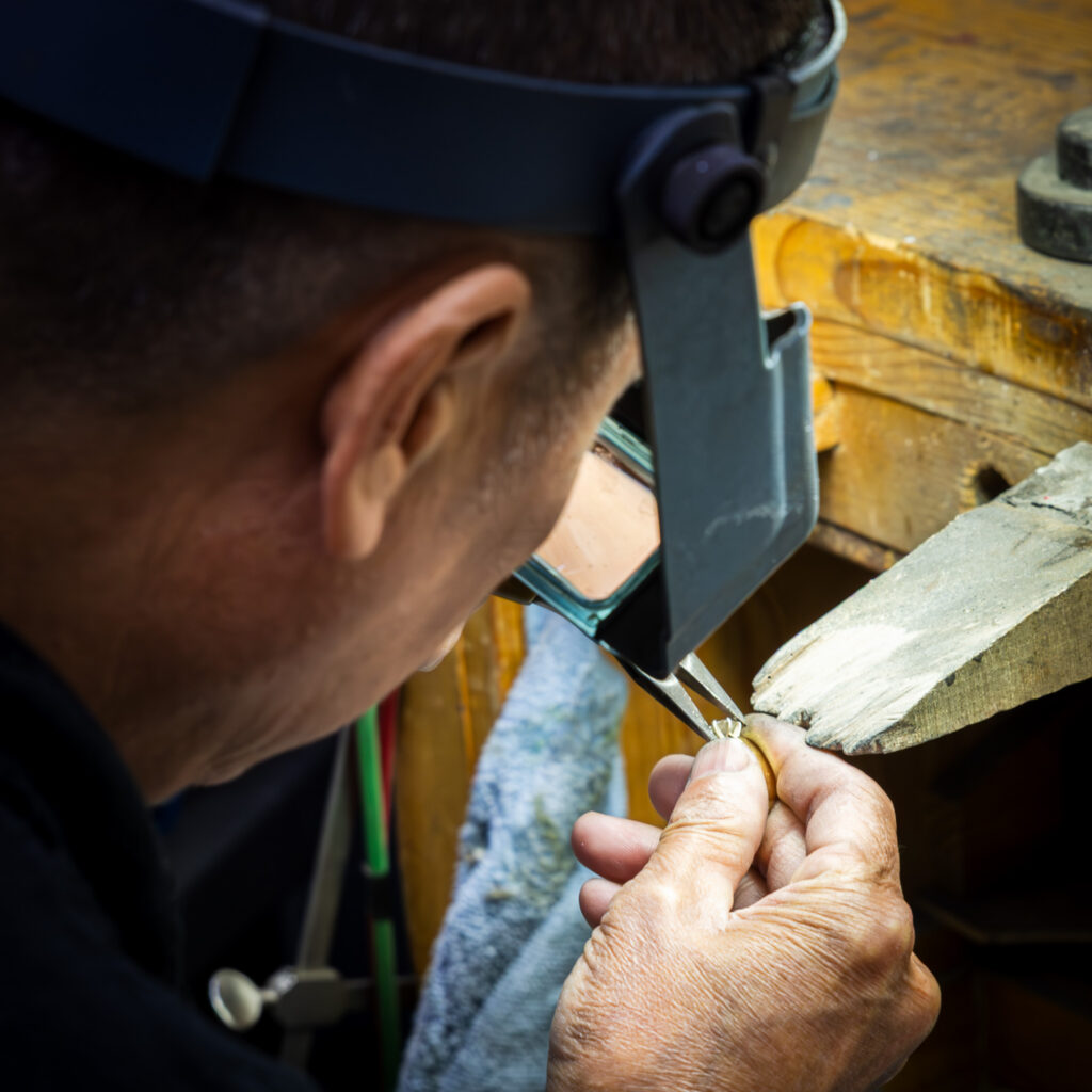 Jeweler wearing a magnifying headband working on a small piece at a wooden bench with tools.