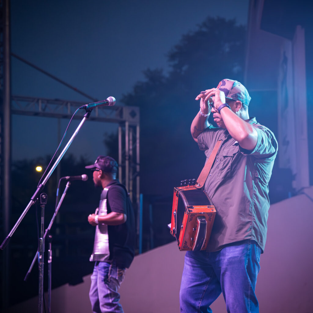 Two musicians performing on an outdoor stage at night, one playing an accordion and the other playing a washboard under stage lights.