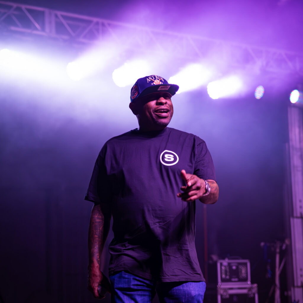Man on stage under purple lighting wearing an Astroworld cap and black T‑shirt, pointing toward the audience during a live performance.