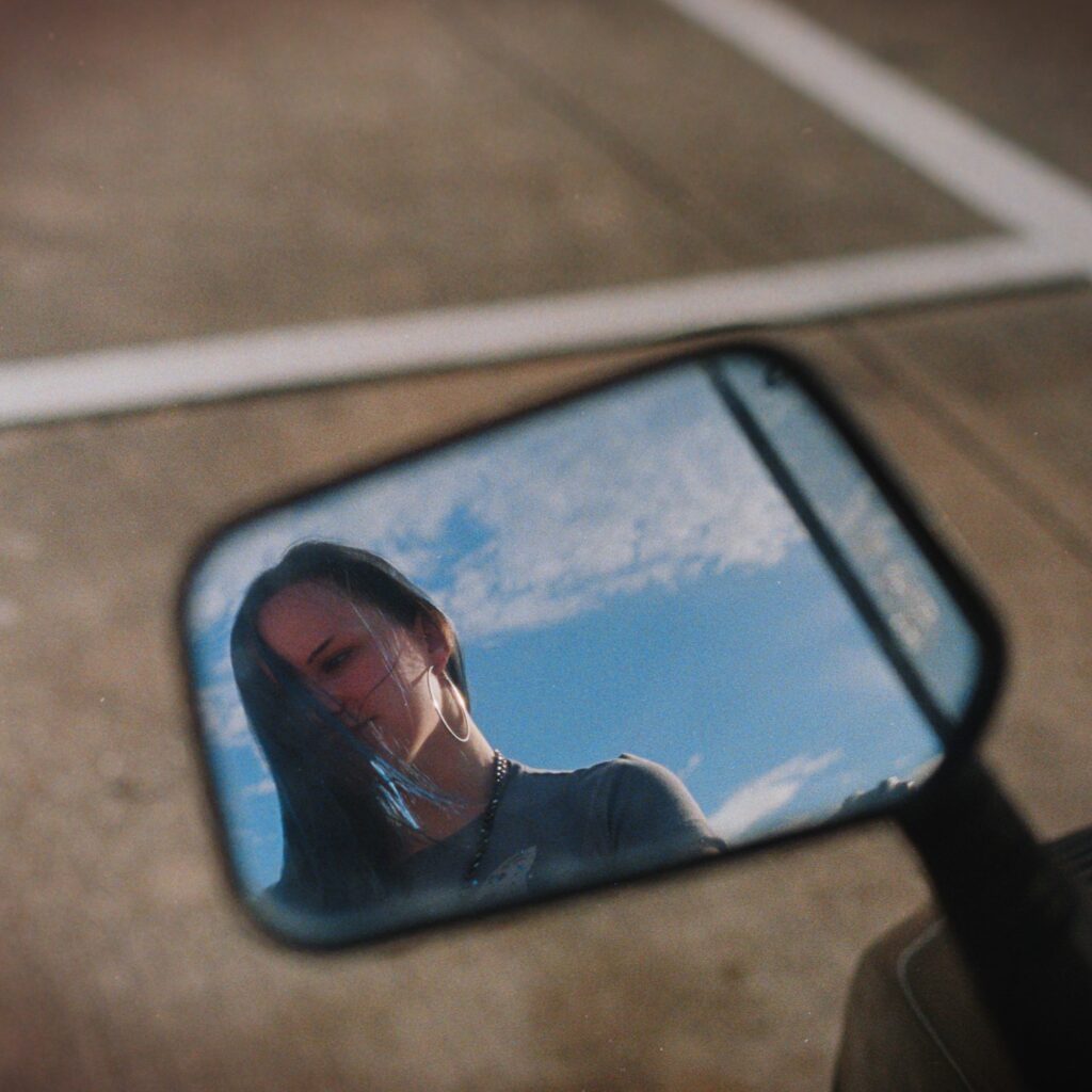 Home Reflection of a person in a motorcycle side mirror against a bright sky in a parking lot.