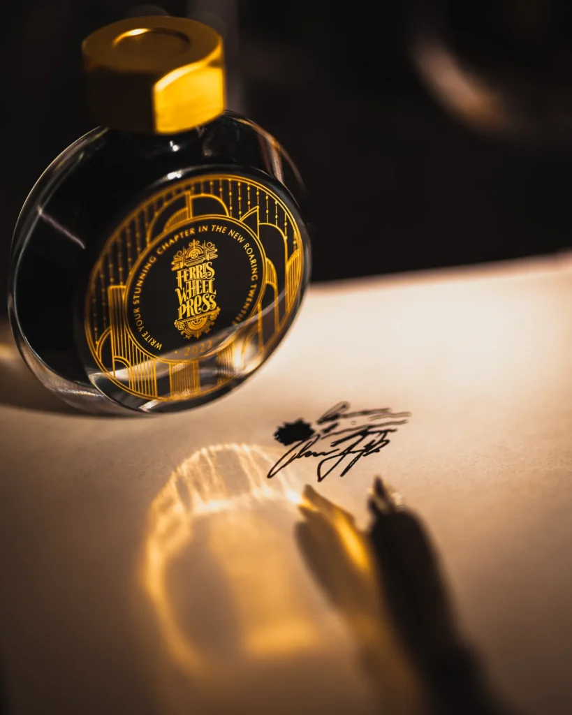 Moody product photo of a small glass ink bottle casting a warm reflection on paper with a pen